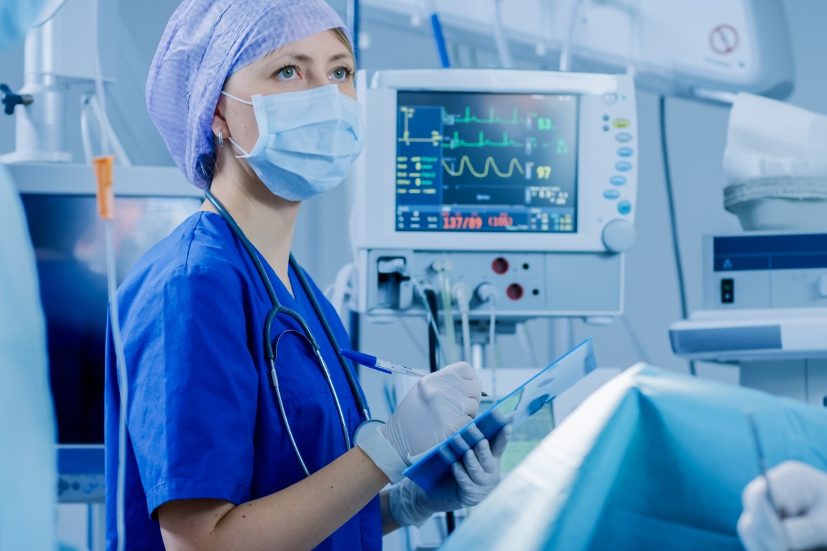 person, wearing hospital scrubs and a mask, writes on a clip board in a surgery room
