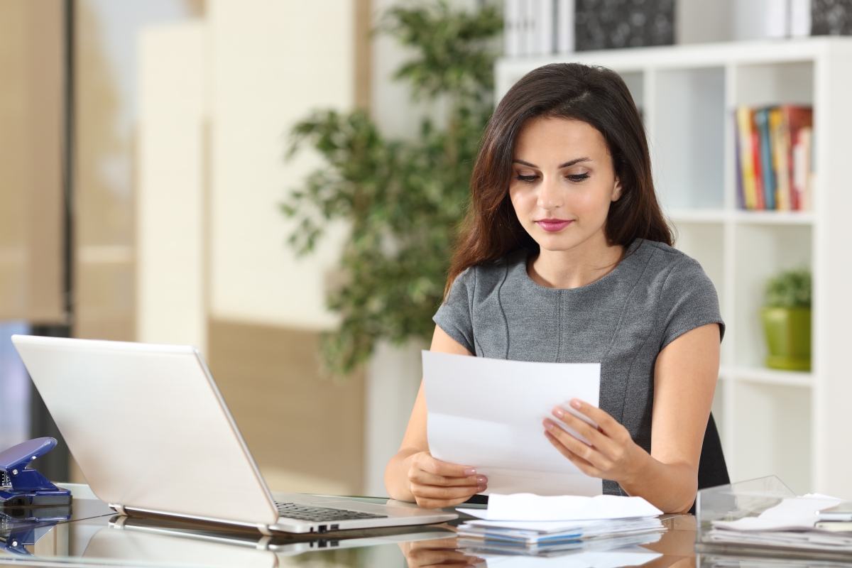 woman reviewing a document while seated at a desk with a laptop in front of her