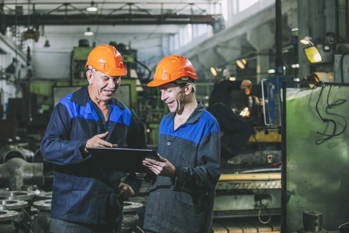 two people, wearing hardhats, looking at a tablet in front of an assembly line