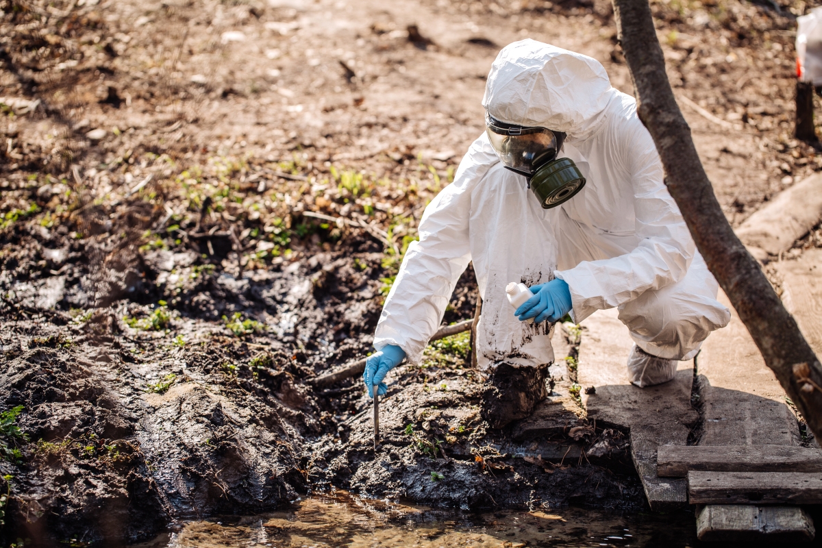 person in a hazmat suit and mask crouching down to collect a sample from the ground