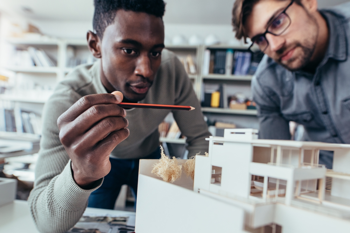 two diverse young men looking at a building model while one gestures with a pencil