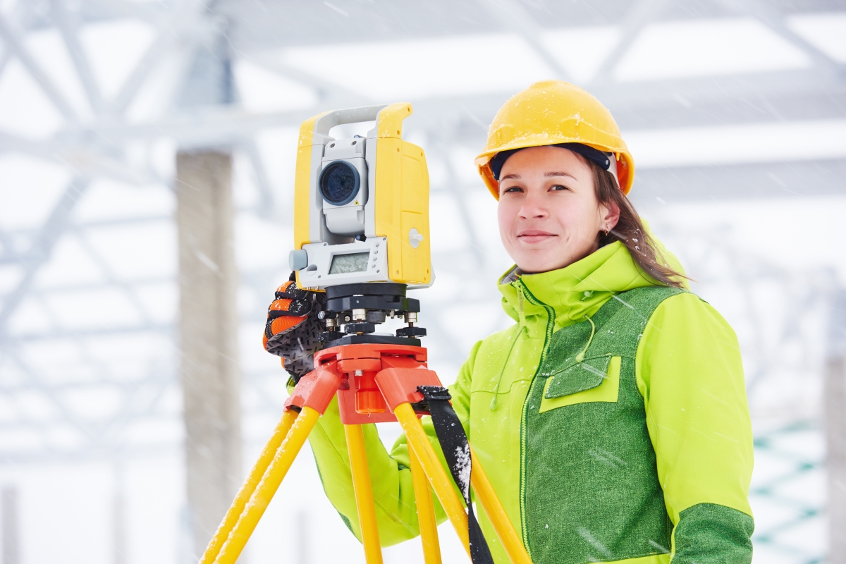 person wearing a yellow hard hat and yellow jacket standing next to a land surveying instrument on a tripod