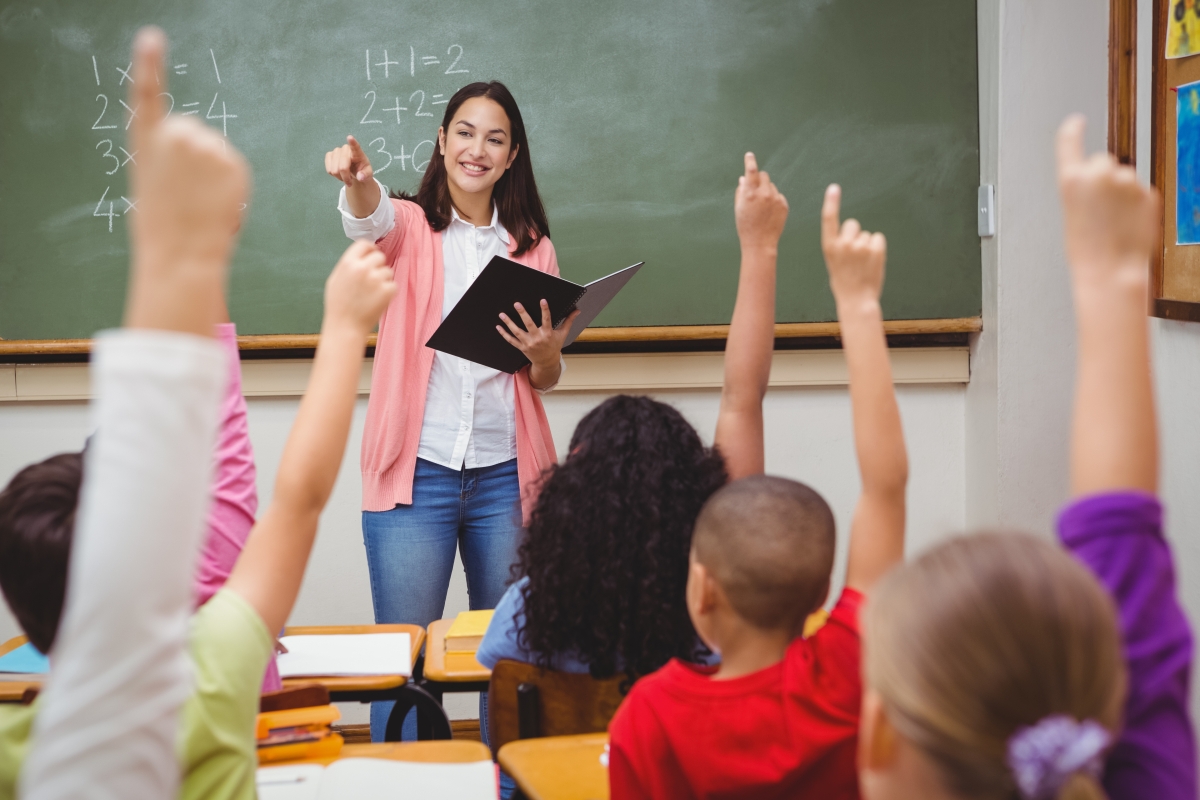 woman standing in front of a chalkboard at the front of a classroom of children with raised hands, pointing at one of the children in the distance