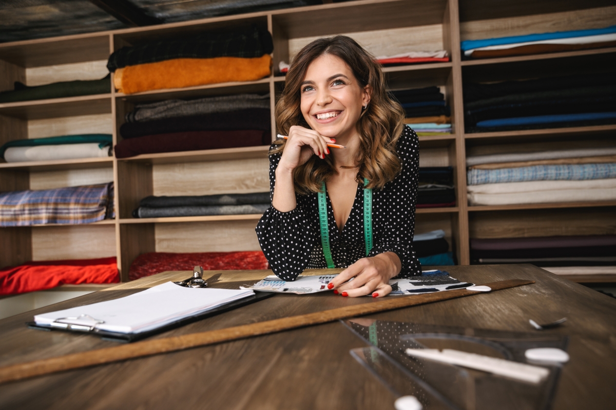 person, leaning on a table with rulers and paper, in front of shelves of fabric