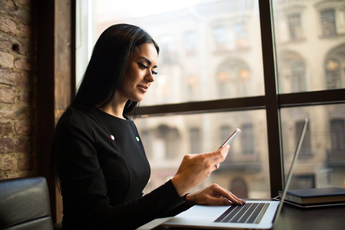 young woman sitting at a table next to a window using a laptop and a smartphone