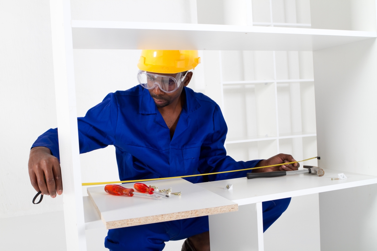 person, wearing a hard hat and safety goggles, measuring and installing a shelf
