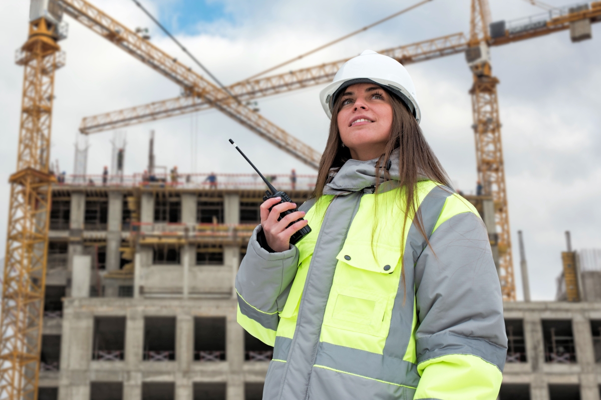 young woman in hard hat and reflective safety jacket holding a walkie talkie at a construction site