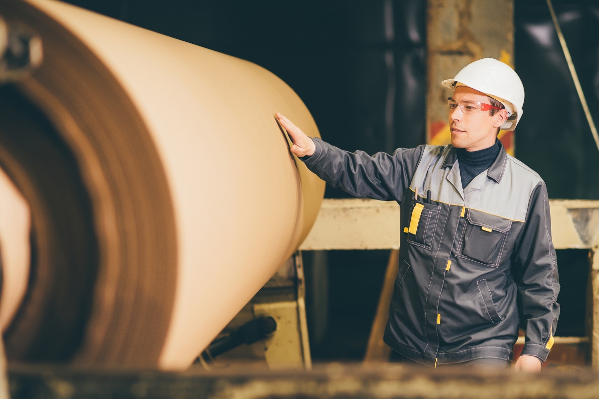 person, wearing a hard hat, inspecting a large roll of brown paper