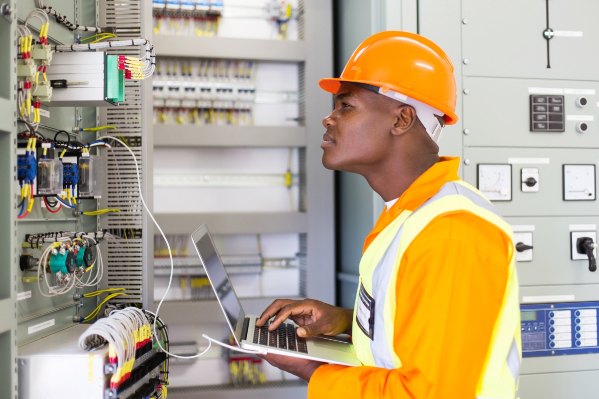 man wearing hard hat and safety attire using a laptop and inspecting electrical equipment