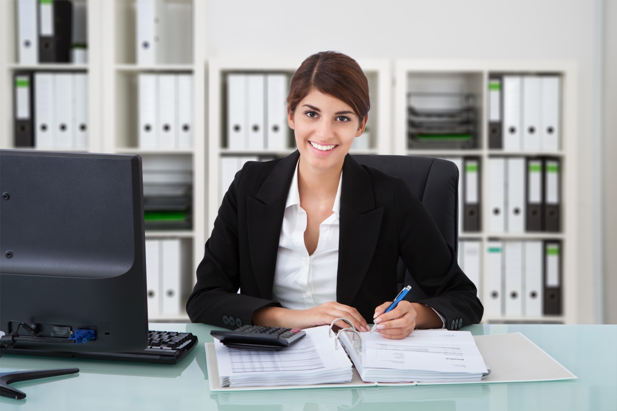 woman wearing business attire sitting at a desk with a computer, calculator and binder open in front of her as she holds a pen