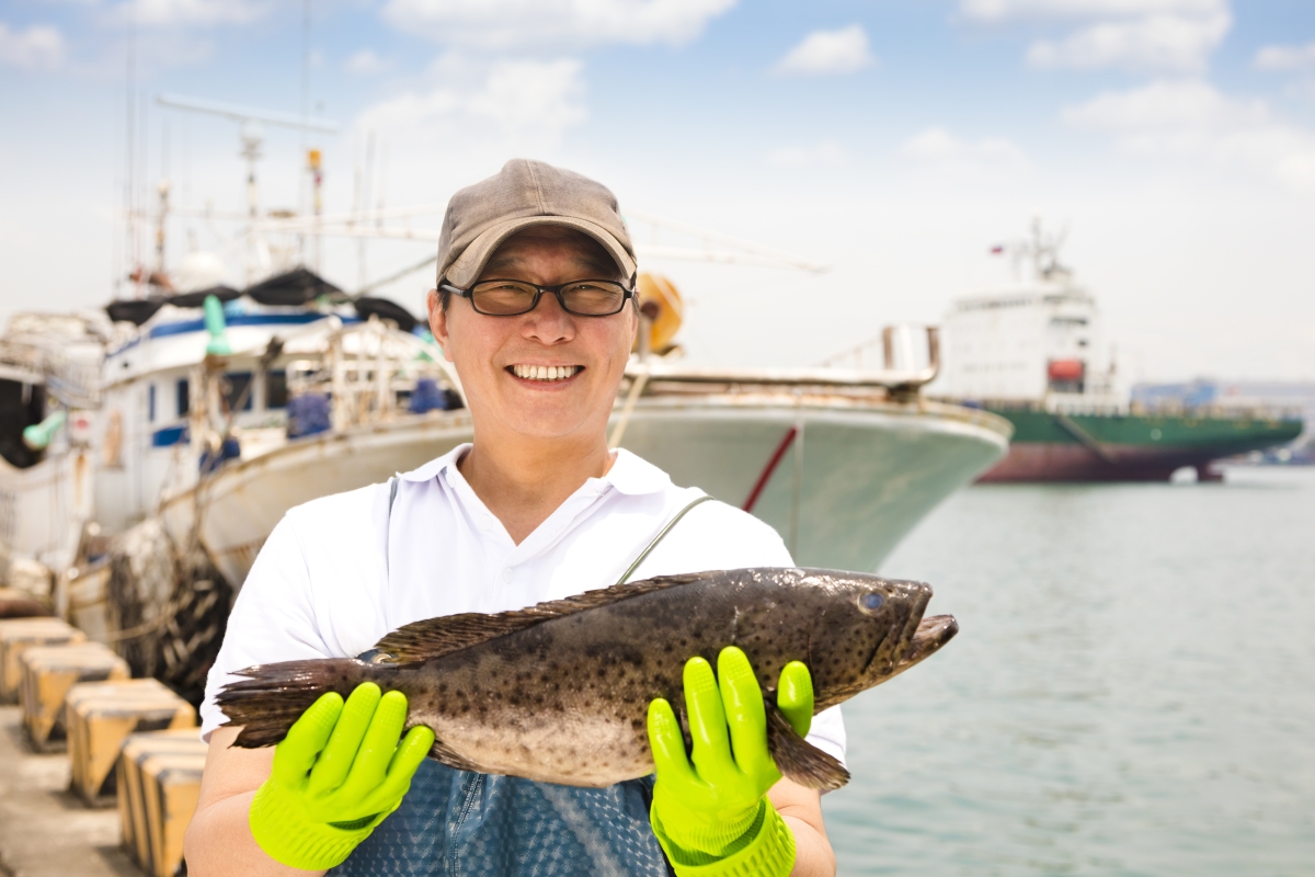 person, wearing gloves, standing in front of a boat and holding a freshly-caught fish