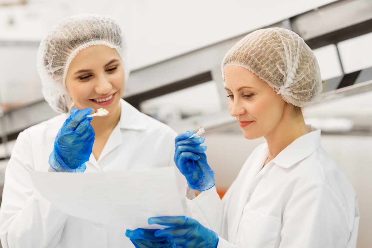 two people, wearing hair nets and gloves, sampling food from plastic spoons