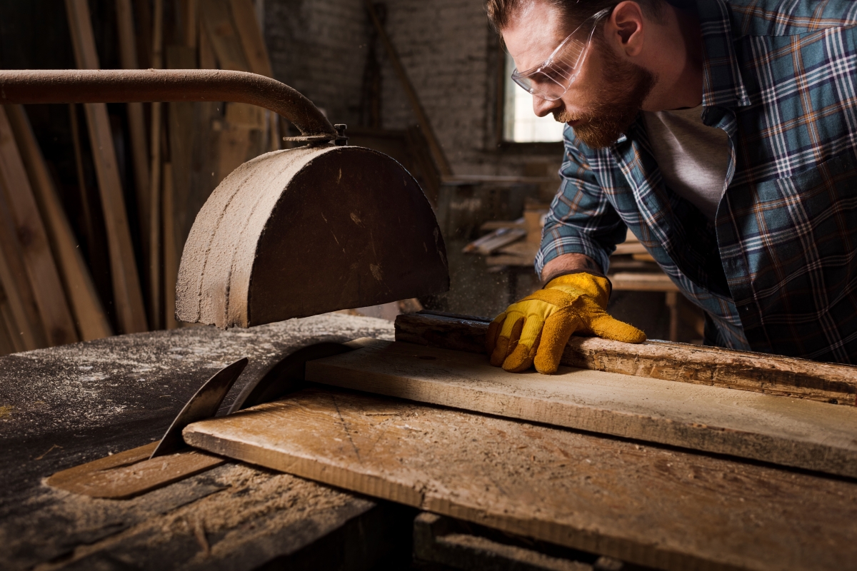 person, wearing safety goggles and gloves, running timber through a saw