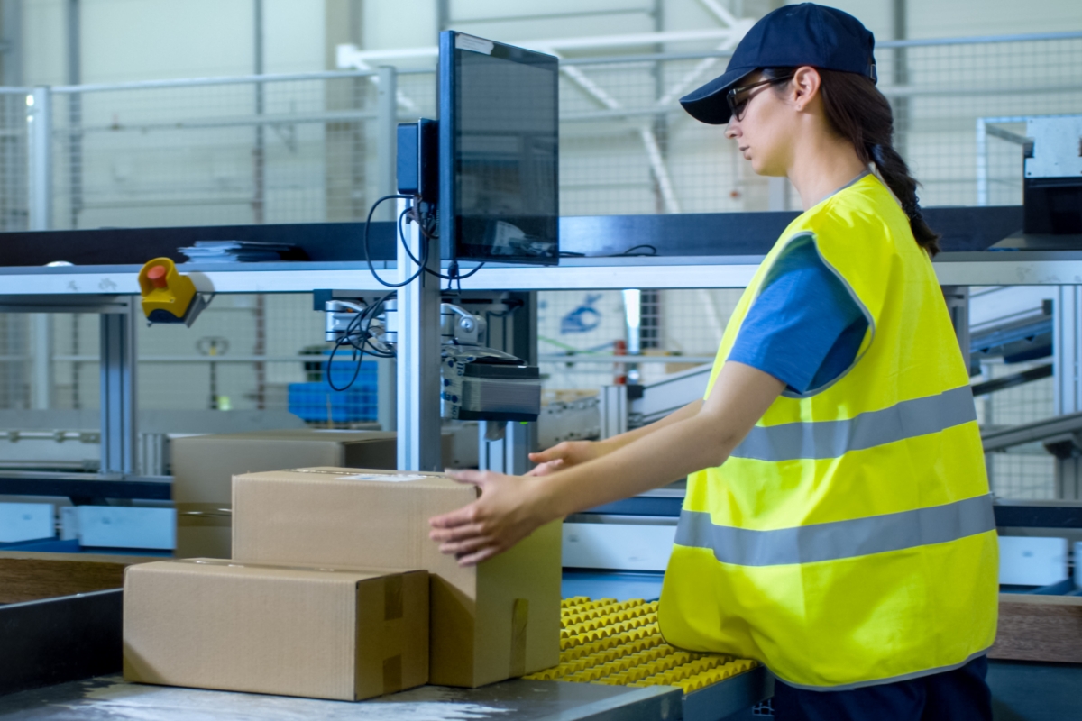 woman wearing a baseball cap and bright yellow safety vest handling boxes in a warehouse while looking at a computer screen