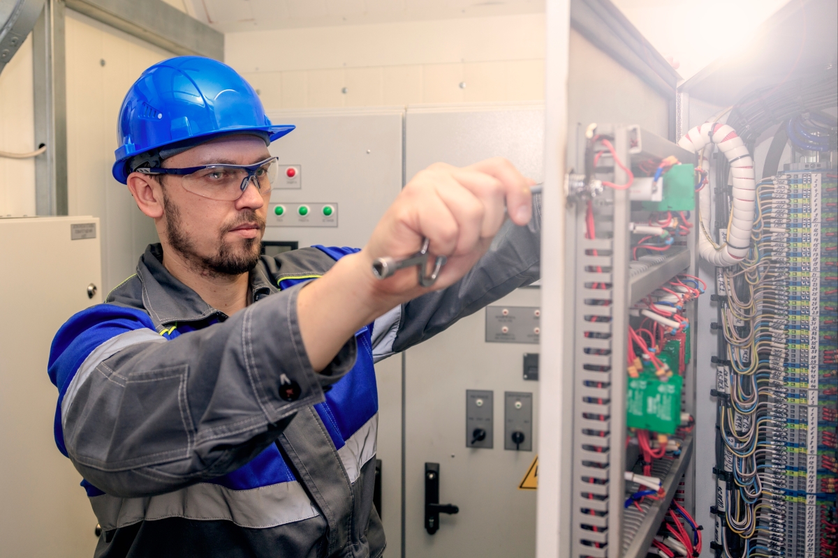 person, wearing a hard hat and safety goggles, working on an electrical panel
