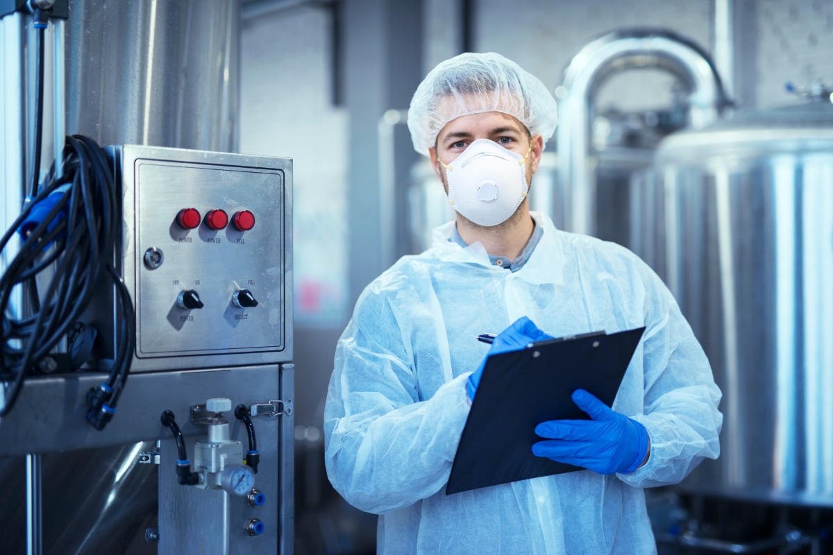 person, wearing a hair net, mask, gloves and plastic coat making a note on a clip board in an industrial environment