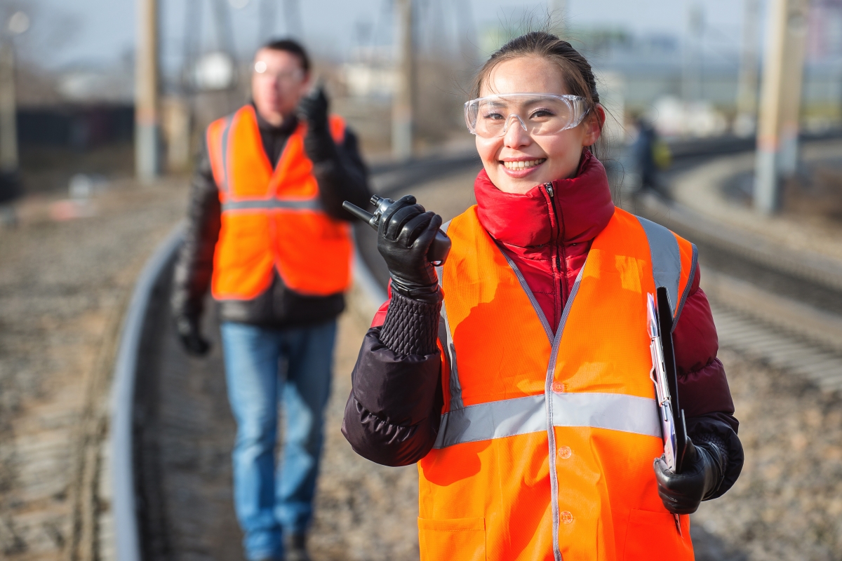woman and man wearing safety glasses and safety vests holding handheld radios while walking along train tracks