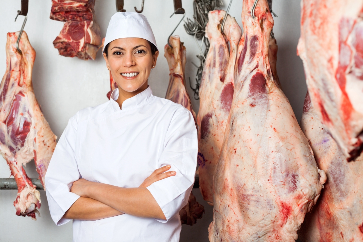 person, wearing a white hat and shirt uniform, standing in front of hanging, large cuts of meat