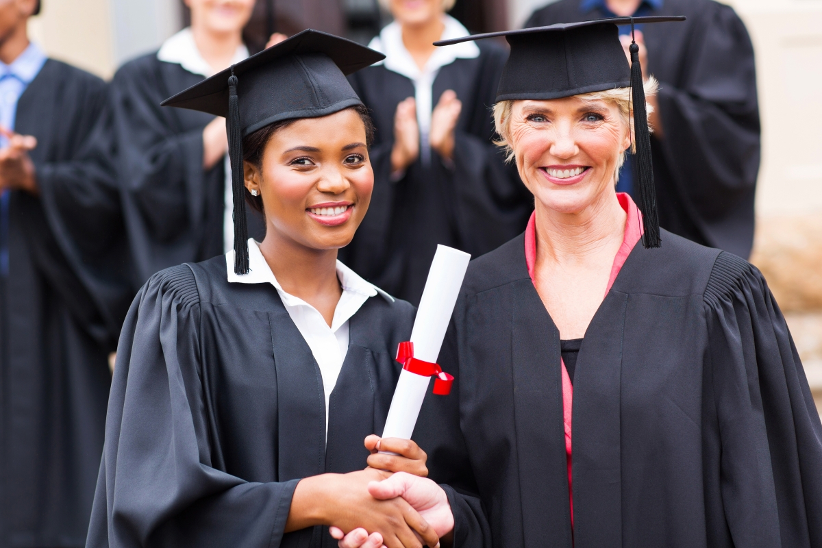 two diverse women both dressed in black graduation caps and gowns smiling and shaking hands while the younger woman holds a diploma