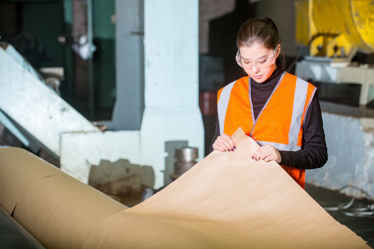 person, wearing safety goggles, inspecting brown paper on a large roll