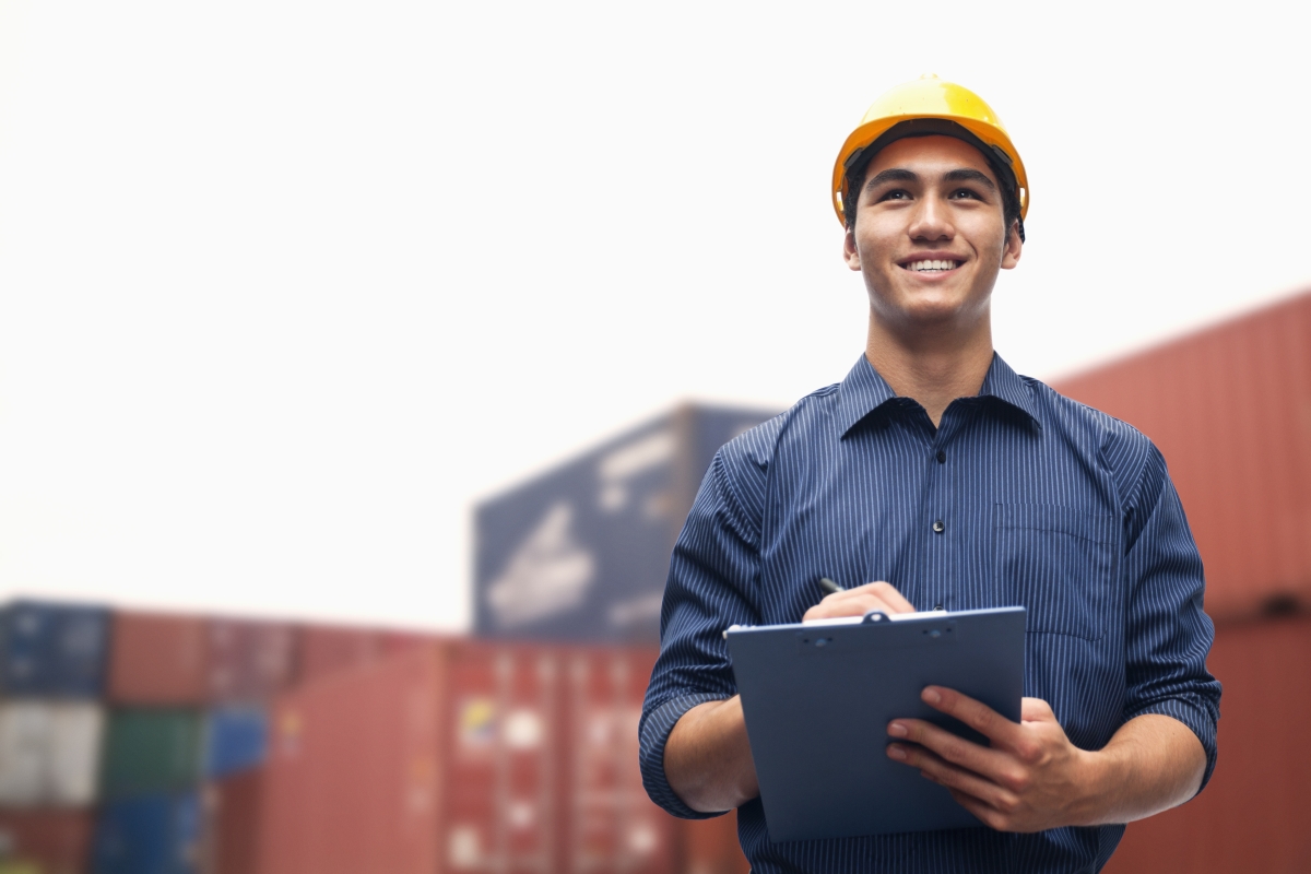 man wearing yellow hard hat writing on a clipboard while standing in front of cargo containers