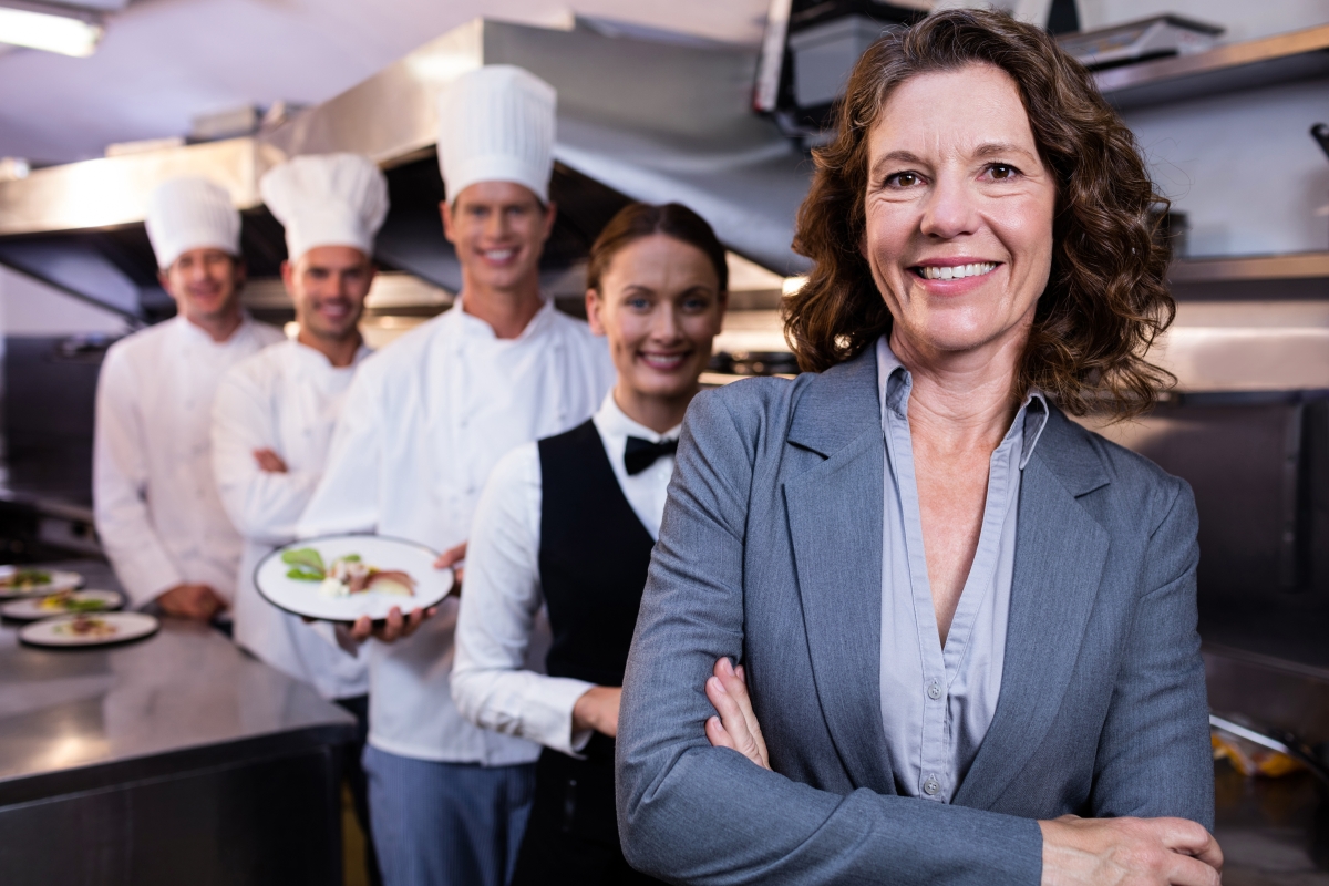 woman in professional attire standing in a kitchen smiling and crossing her arms with three chefs and a server standing behind her also smiling