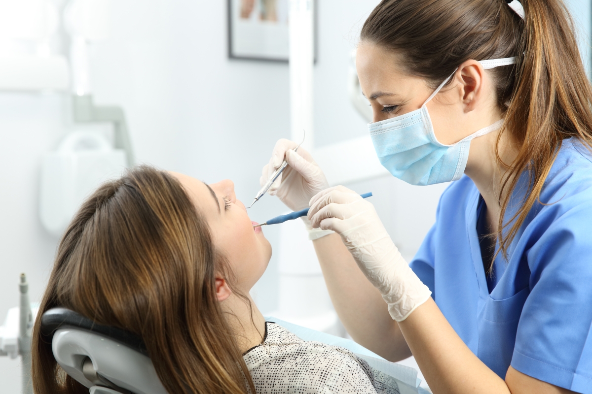 woman wearing scrubs and a mask with her hair tied back using dental tools to work on a patient's teeth