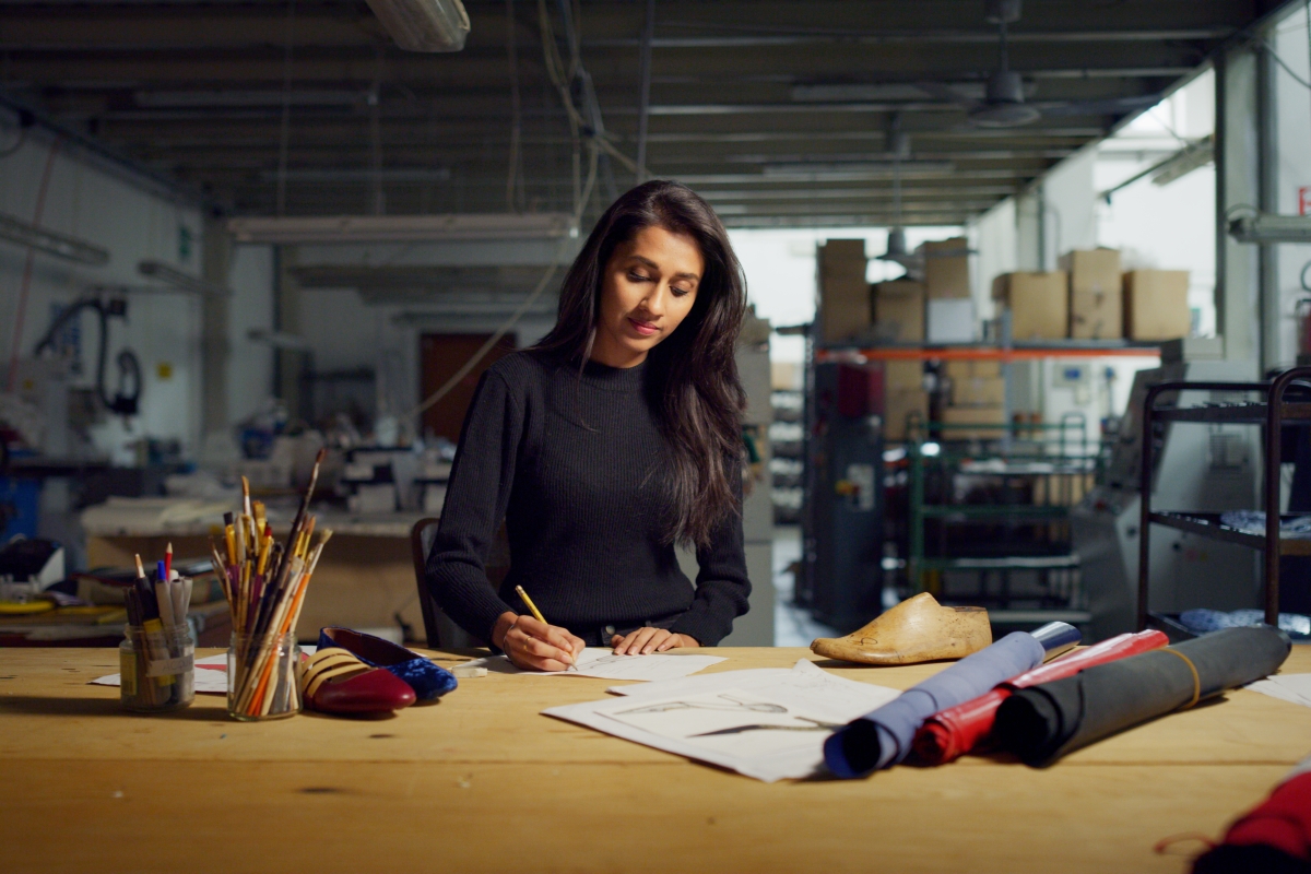 young woman sitting at a large wooden table with materials and art supplies on it while drawing designs on paper